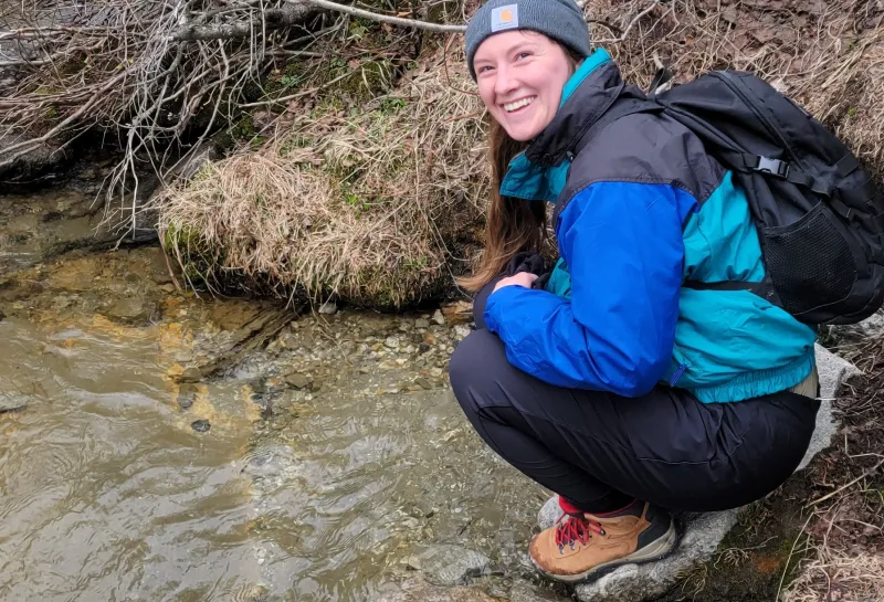 Molly crouched by a creek wearing hiking gear and a backpack.