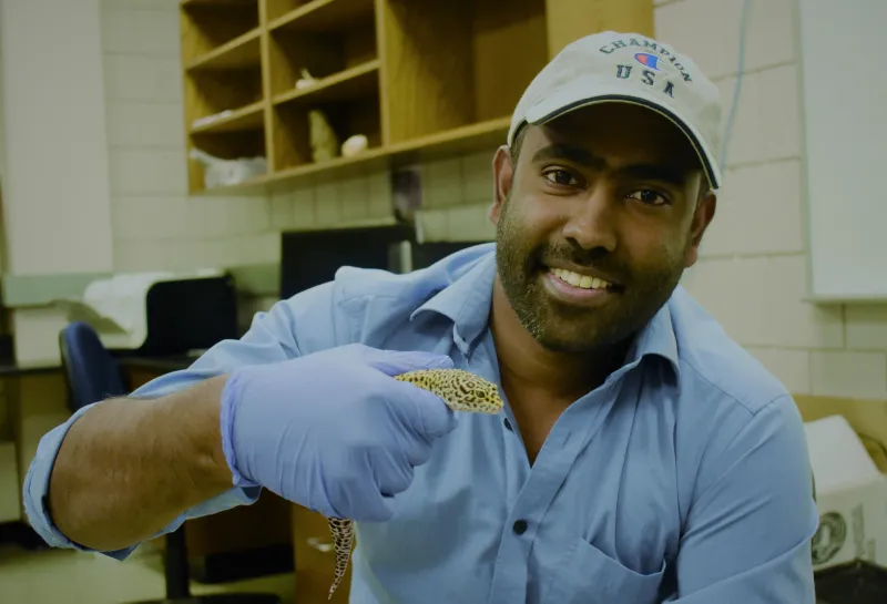 Sekhar smiling confidently at camera while holding an animal with blue gloves.