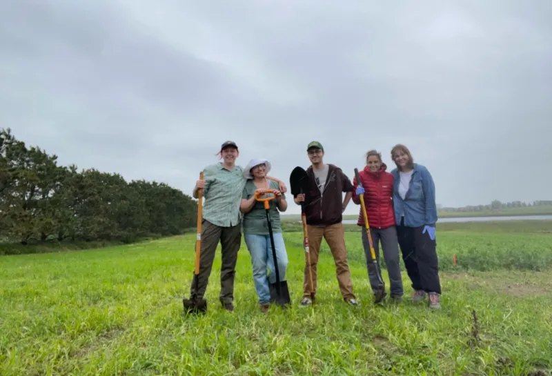 Students standing in field holding shovels and smiling confidently at camera.