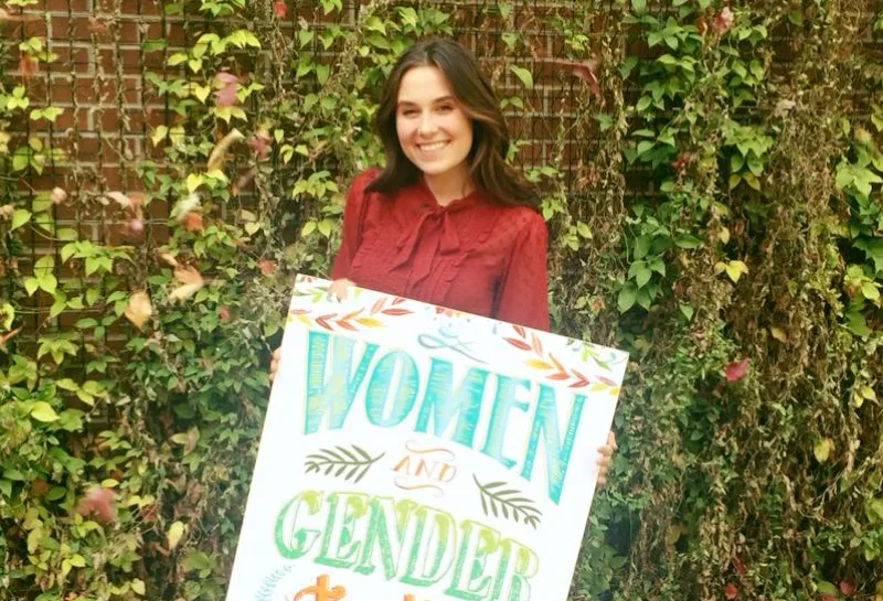 Woman holding a Women and Gender studies sign standing in front of greenry on a brick wall.
