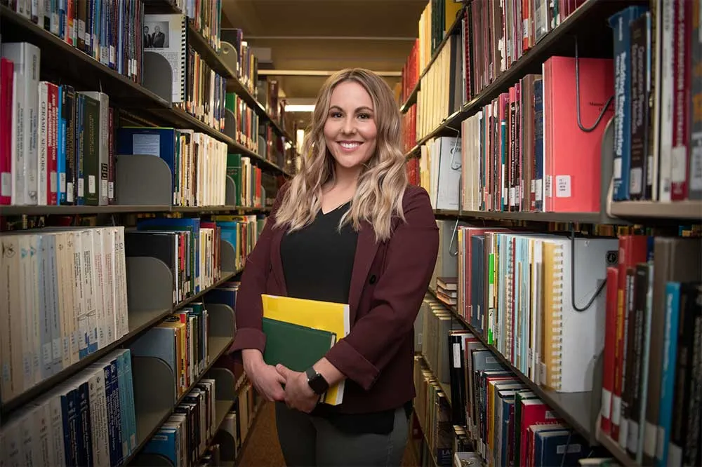 Student standing is aisle between two rows of bookshelves in the library.