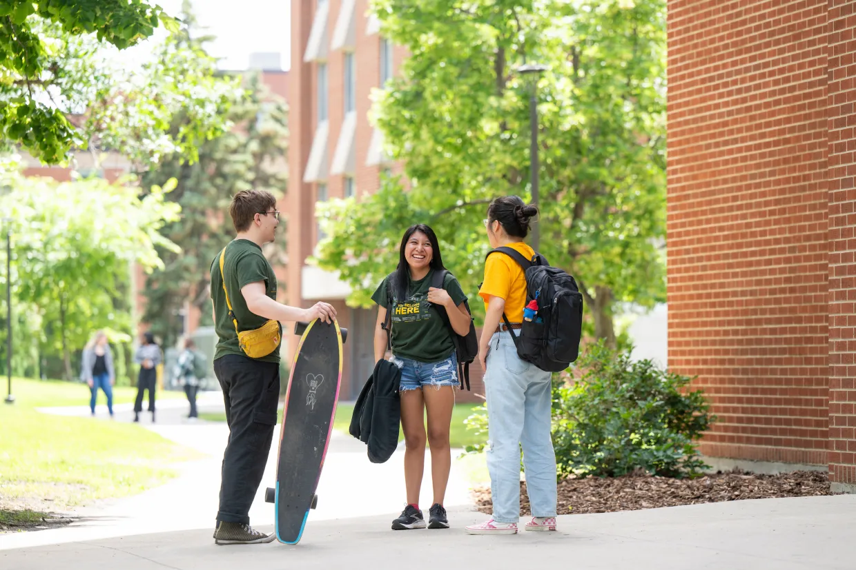 Three students chatting on campus wearing backpacks. The lefthand student has a longboard skateboard.