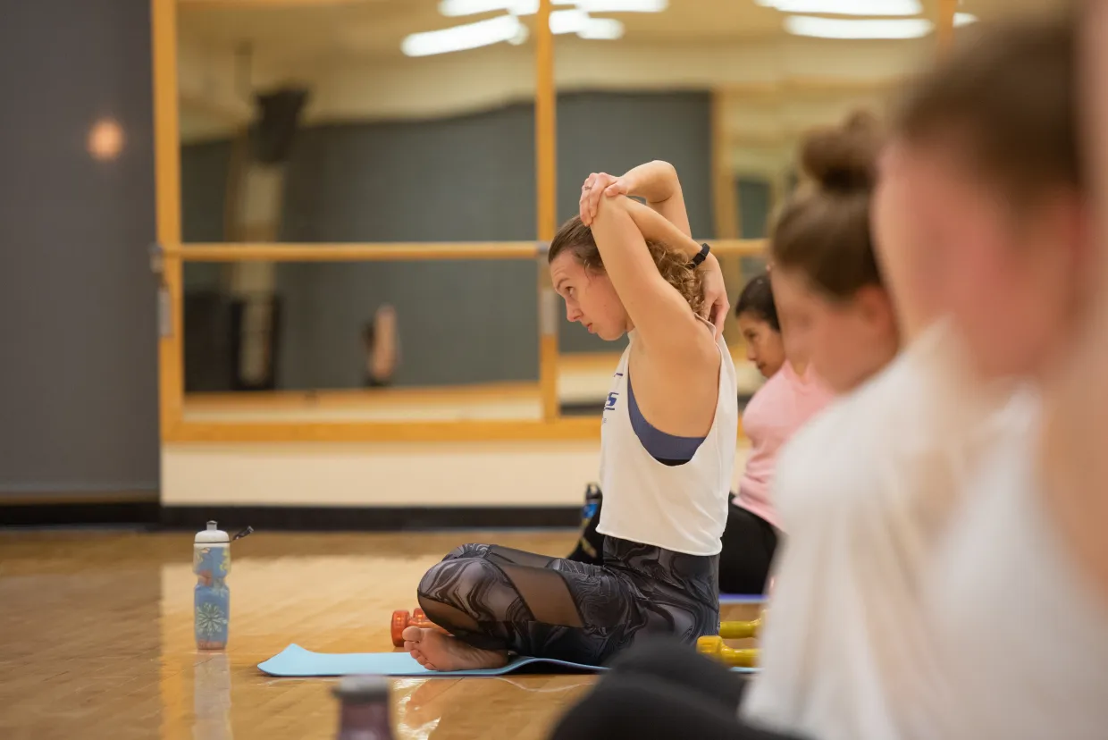 Woman sitting cross legged on a blue yoga mat wearing a white tank top and black leggings. She is stretching her arms in front of a mirror.