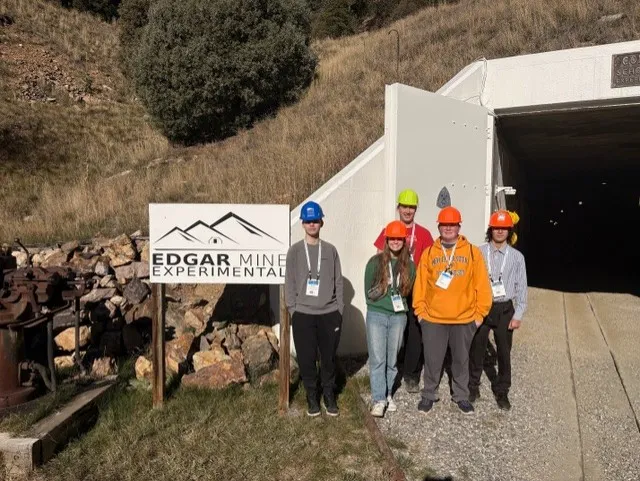 Physics students in hard hats stand by the opening to a mine.  Sign to the side reads "Edgar Mine Experimental"