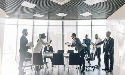 Business people around a conference room table with two people arguing and the rest standing around the table