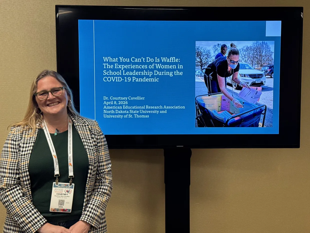 Presenter standing in front of TV display of research presentation title and presenter information.