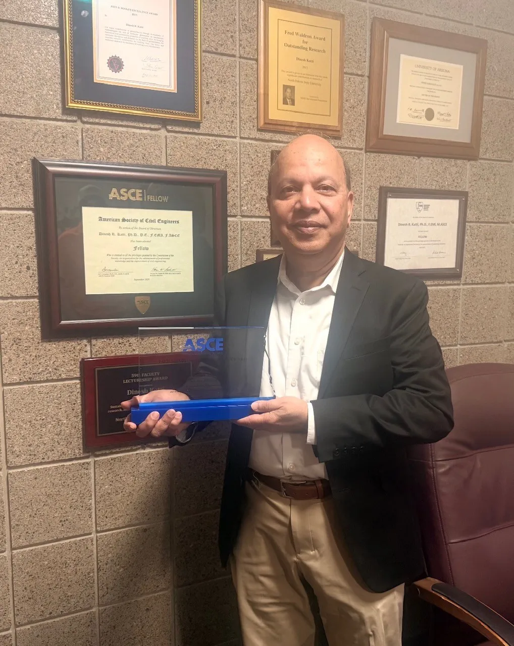 Dr. Dinesh Katti holds his ASCE award in his office near other awards against the wall.