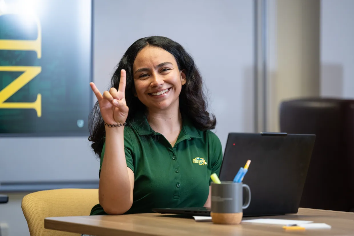 a female student in a green NDSU polo sits in front of a laptop with her hand in a 'Horns up' sign.