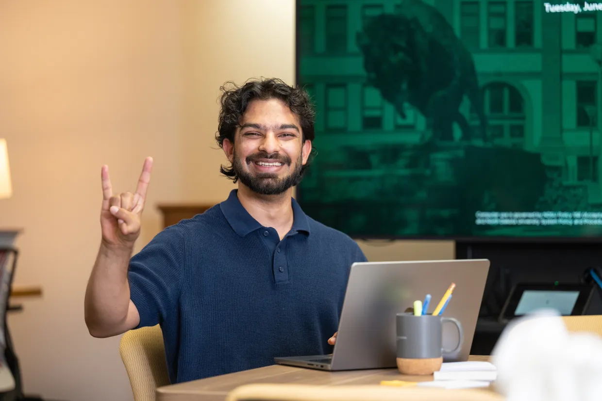 A male student in a blue polo sits in front of a laptop with his hand in a 'Horns up' sign.
