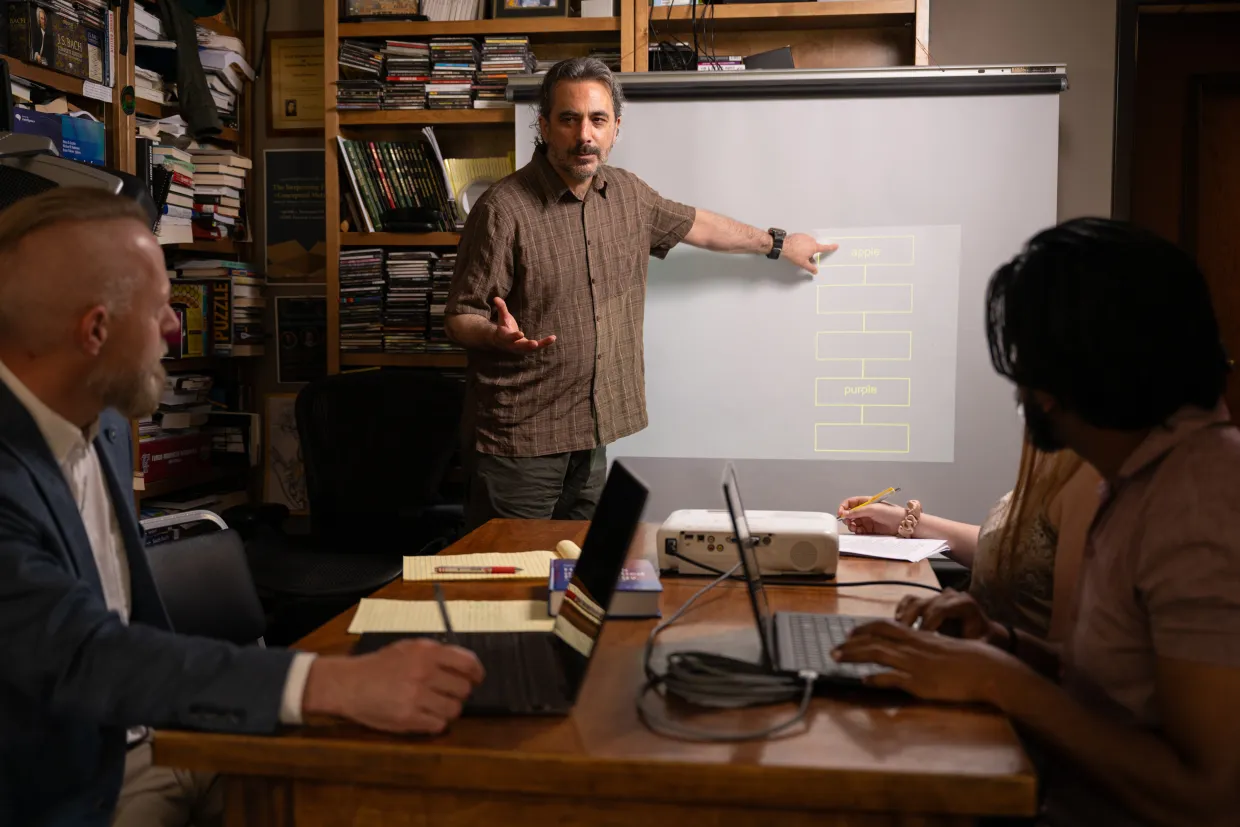 University Distinguished Professor of psychology, Michael D. Robinson, points at a whiteboard while students sit around a desk on their laptops.