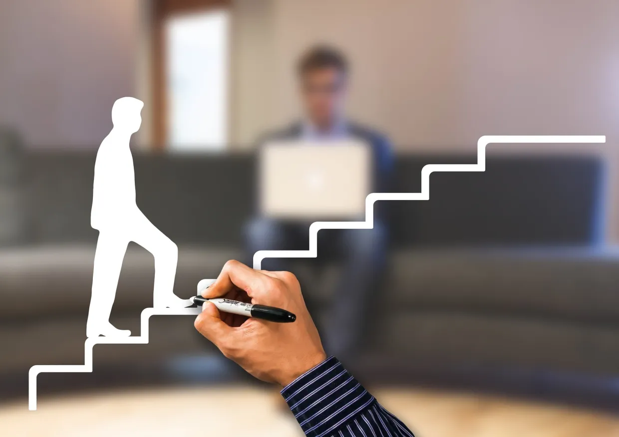 Person drawing a person climbing stairs on a clear board with a young man on a laptop in the background