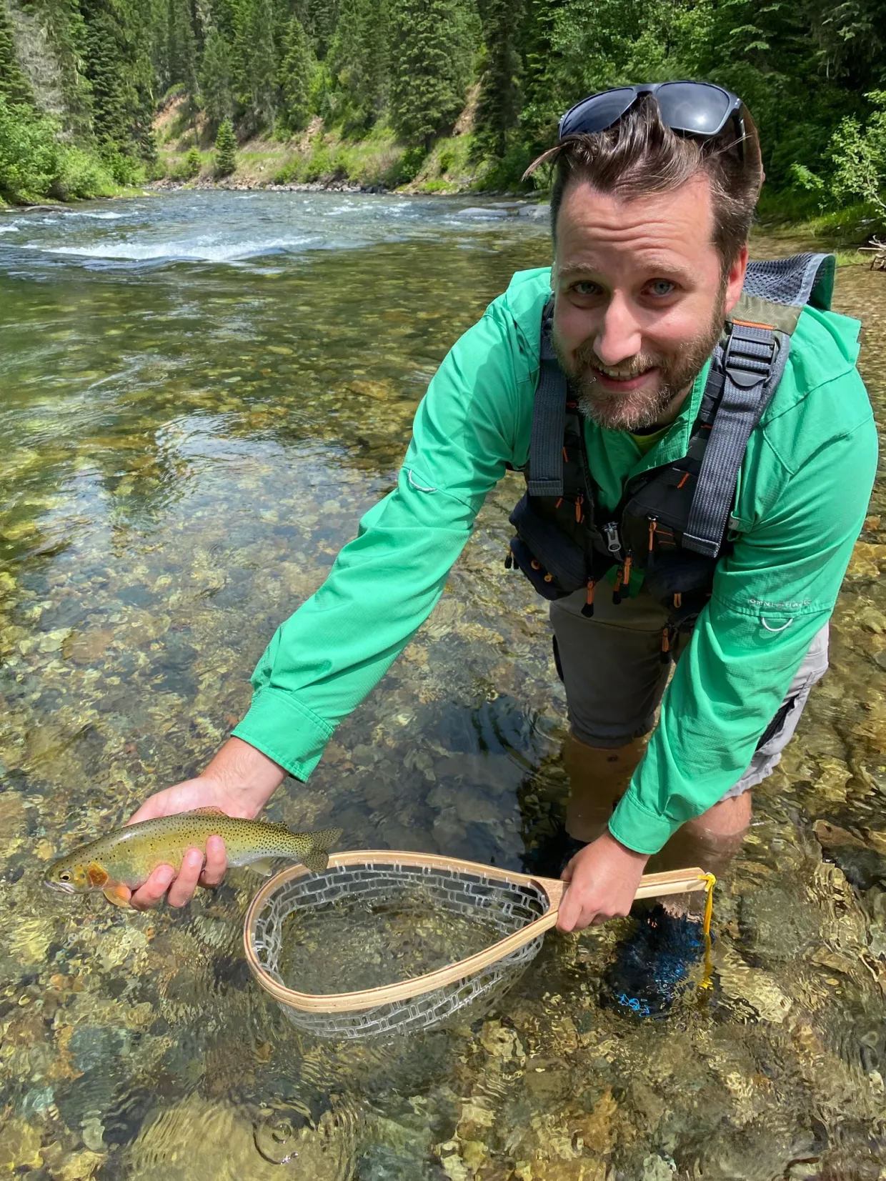 Travis standing in a river holding a net and fish while smiling at camera. 