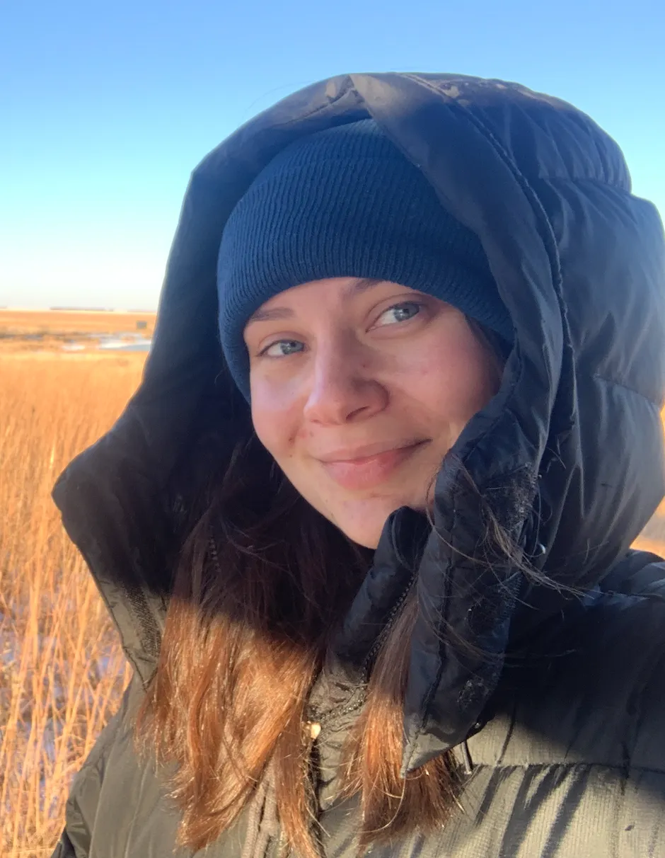 Josie Pickar standing in field wearing jacket with hood over her head while looking confidently at camera.