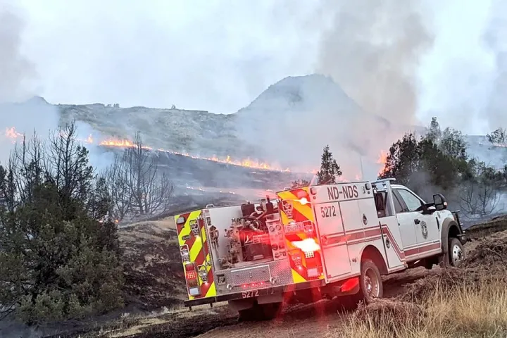 White fire engine with flashing red lights parked near a hill that is on fire with smoke trailing up into the sky