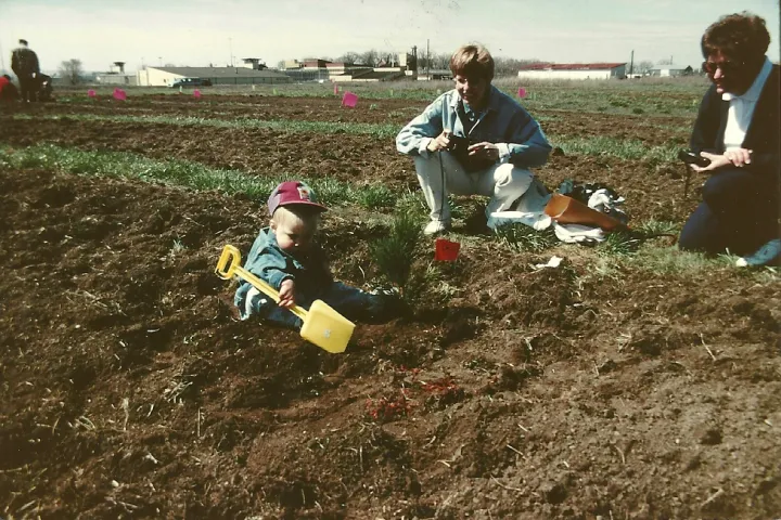 A young child uses a yellow hand shovel to plant a tree