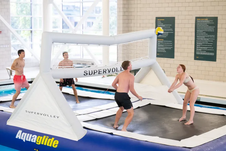 Three males in black swim trunks and one woman in a white swim suit playing volleyball on a floating trampoline in Wellness Center lap pool
