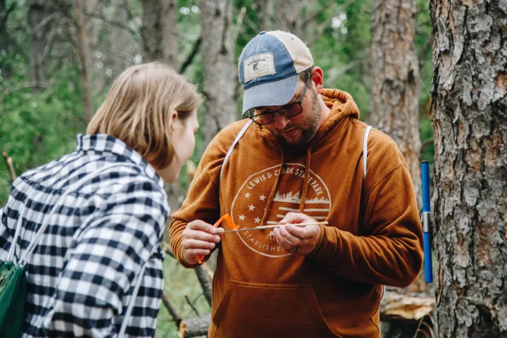 A man in a hoodie and ballcap looks at a tree core with a young woman in a flannel