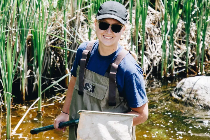 A woman in waders, a baseball cap, and sunglasses smiles while holding a net and stands in a river lined with cattails
