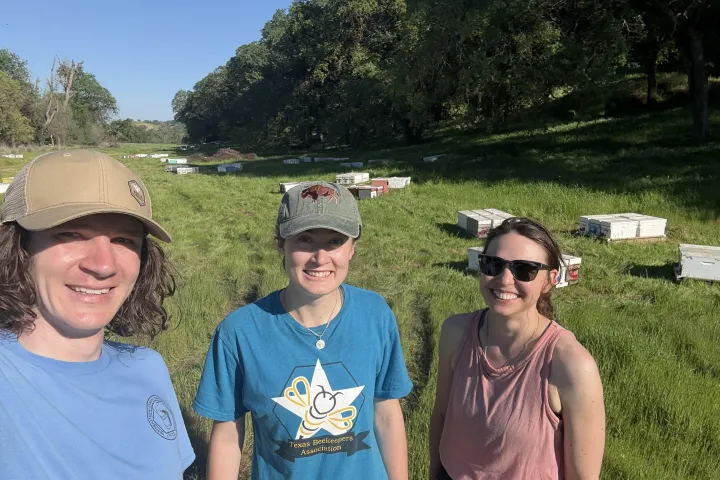 NDSU assistant professor Rogan Tokach standing on grass with two students along with scattered beekeeping equipment in the background.