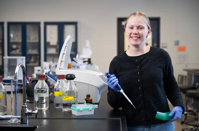 Nora standing in lab with research materials, smiling at camera