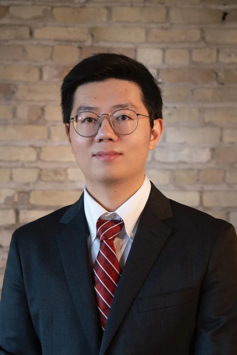 A man with black hair standing in front of a brick wall wearing a black suit and red striped tie, and wire frame glasses.