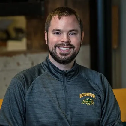 A man with short brown hair and a beard smiling, wearing a gray NDSU quarter zip shirt