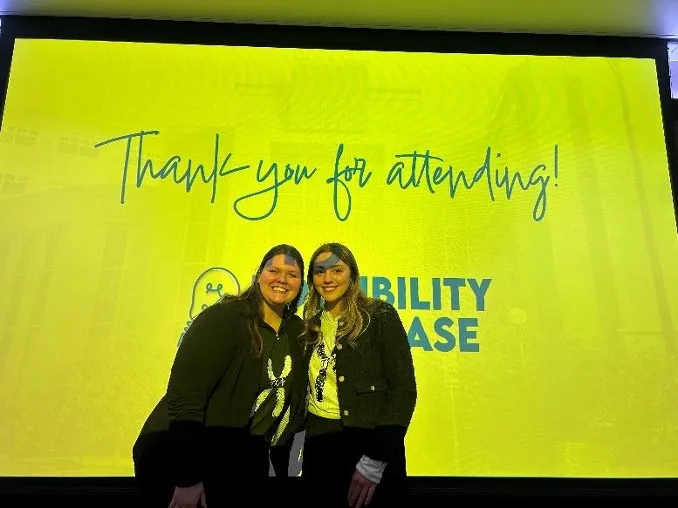 Two student smiling confidently at camera while posing in front of yellow screen.