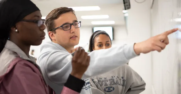 Three students in a classroom looking at information on a whiteboard. One student is pointing at the content on the board. 