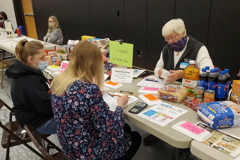 An older adult sitting across a table from two teens. The table is covered in grocery items, a calculator and other papers. There is a handwritten sign that read "Great Groceries".