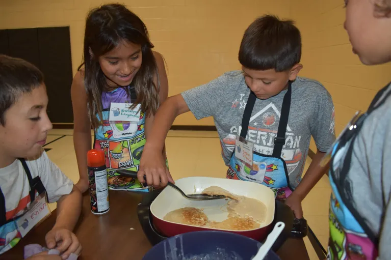 Kids gathered around an electric frying pan cooking pancakes